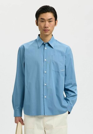 Homme portant une chemise bleu clair à manches longues boutonnée et un pantalon blanc, debout avec une main dans la poche et tenant un objet beige.