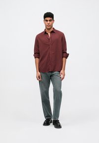 Vīrietis, kurš valkā garam piedurknēm bordo kreklu, pelēkas džinsus un melnas kurpes, stāv uz neitrālas baltas fona.