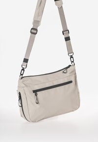 Borsa a spalla in tessuto beige con tracolla regolabile, due tasche con zipper e dettagli in hardware nero. Textura liscia e forma rettangolare.