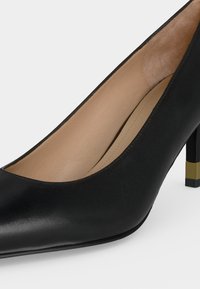 Gros plan d'une chaussure à talons hauts en cuir noir avec un bout pointu et un accent doré sur le talon sur un fond uni.