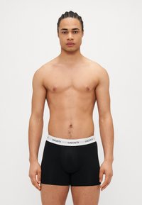 Fekete boxer alsónadrág fehér gumírozott derékkal, amelyen a 'Lacoste' logó található. Az anyag simának tűnik, látható minták vagy díszítések nélkül.