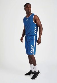 Niebieski sportowy top bez rękawów z białą lamówką, w zestawie z dopasowanymi niebieskimi szortami z białymi akcentami w kształcie szewronu po bokach. Noszone z czarnymi tenisówkami.