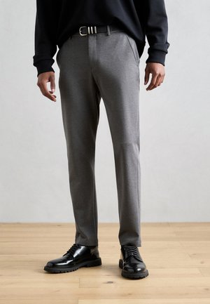 Pantalones grises a medida hechos de una tela suave, con un corte ajustado, bolsillos laterales y un cinturón oscuro. Combinados con zapatos de vestir negros brillantes.