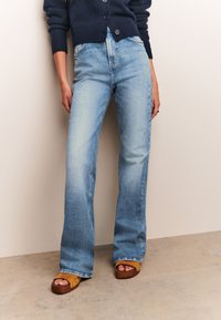 Šviesiai mėlynos platėjančios džinsinės kelnės pasižymi išblukusiu plovimu, klasikiniu penkių kišenių stiliumi ir siūlių detalėmis. Derinamos su rudos spalvos platforminiais sandalais.