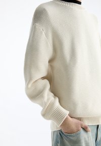 Persoon in een crème kleurige, gestructureerde gebreide trui met ribbels aan de mouwen, met één hand in de zak van een lichtblauwe spijkerbroek.