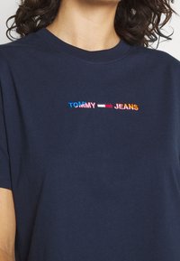 Camiseta de algodón azul marino con el texto bordado "TOMMY" en azul y "JEANS" en amarillo, con un acento rojo y blanco en el medio.