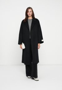 Black long coat with wide sleeves, paired with a gray top and wide black pants. Accessorized with a small white handbag and light shoes.