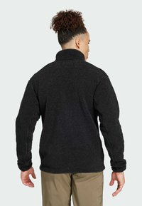 Pullover in pile nero, colletto alto, superficie testurizzata, maniche lunghe, vestibilità rilassata, visto da dietro con le mani ai fianchi, abbinato a pantaloni khaki.
