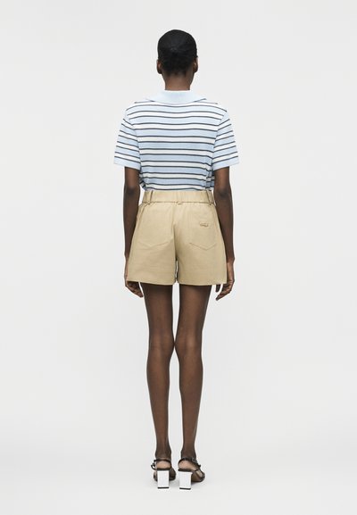 Άτομο που στέκεται με την πλάτη, φοράει μπλε και λευκή ριγέ κοντομάνικη μπλούζα, μπεζ ψηλόμεσο σορτς και μαύρα σανδάλια με τακούνι.