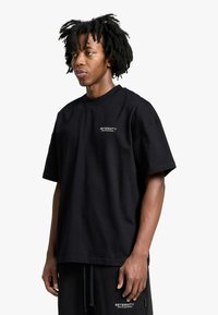 T-shirt nera in cotone con vestibilità rilassata, maniche corte e scollo arrotondato. Presenta un piccolo logo bianco sul petto e una trama visibile.