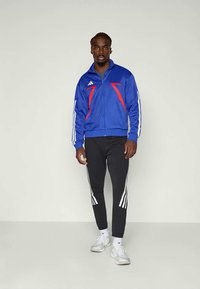 Blå sportsjakke med røde detaljer, hvite striper på ermene; svarte bukser med hvite striper; modell iført hvite sko, stående oppreist.