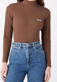 Top de manga larga de canalé marrón con cuello alto, que presenta un pequeño logo blanco. Combinado con unos jeans de mezclilla de tiro alto en azul claro.