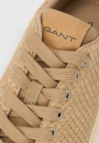 Κοντινό πλάνο μπεζ υφασμένο αθλητικού παπουτσιού με κορδόνια στο ίδιο χρώμα και ετικέτα γλώσσας από ταμπά δέρμα που δείχνει το όνομα της μάρκας "GANT".