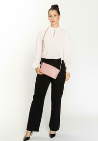 Borsa a tracolla rosa con finitura testurizzata, forma slim e tracolla. Il modello indossa una blusa bianca e pantaloni neri, in piedi contro uno sfondo bianco.