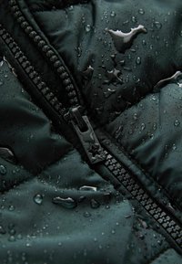 Vandafvisende mørkegrøn pufferjakke med en glansfuld sort lynlås og vanddråber på en struktureret overflade.