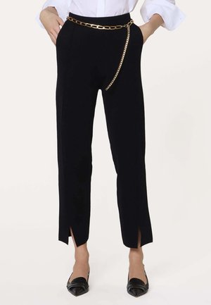 Zwarte op maat gemaakte enkellange broek met voorste splitten, gecombineerd met een gouden kettingriem en zwarte puntige loafers, gedragen met een wit overhemd.