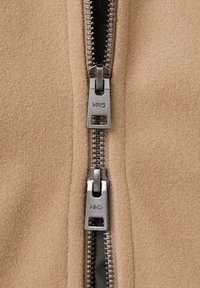 Beige jakke i uldblanding med sort lynlås og sølvfarvet hardware. Synlige lynlåstænder og mærkenavn præget på trækfane.
