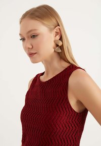 Top senza maniche in maglia bordeaux con un motivo a zigzag testurizzato; presenta grandi orecchini scultorei in tono oro.