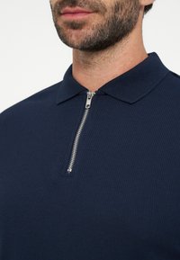 Homme portant un polo bleu marine texturé avec une fermeture éclair argentée à l'avant, montrant le bas du visage et le cou avec une barbe courte.