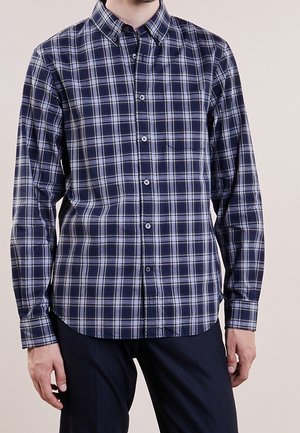 Homme portant une chemise à carreaux bleu marine et blanc à manches longues avec des boutons, et un pantalon foncé, debout devant un fond uni.