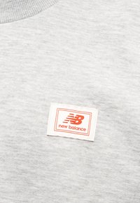 Γκρι φούτερ με ριμπ λαιμόκοψη, απαλή υφή και λευκή ετικέτα με πορτοκαλί λογότυπο και έμβλημα «New Balance».