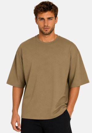 Homme aux cheveux bruns courts et avec des poils au visage, portant un t-shirt marron à manches courtes et ample ainsi qu'un pantalon noir sur fond uni.