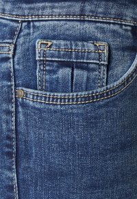 Jeans de denim com uma cor azul média, um bolso traseiro com costura visível e um pequeno detalhe em rebite metálico.