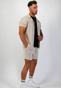 Beige kortärmad skjorta med knappar över en svart t-shirt, tillsammans med beige shorts med dragsko och vita sneakers med svarta ränder.