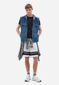 Chaleco de mezclilla con capucha, dos bolsillos frontales, llevado sobre una camiseta negra. Combinado con shorts blancos con rayas negras y una camisa de cuadros atada a la cintura.