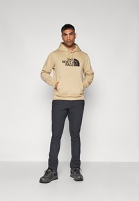 Hanorac bej cu logo negru, buzunar frontal și sfoară de ajustare. Pantaloni întunecați și pantofi gri cu accente galbene. Design simplu și croială relaxată.