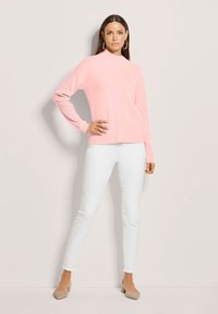 Frau in einem hellrosa Pullover, weißen, figurbetonten Hosen, beigen Ballerinas und großen Creolen, die vor einem schlichten hellen Hintergrund steht.