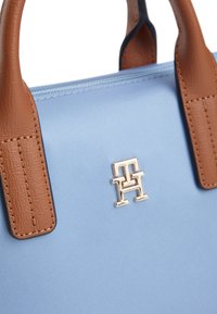 Τσάντα χειρός σε ανοιχτό γαλάζιο χρώμα με καφέ δερμάτινες λαβές και μεταλλικό λογότυπο με τα γράμματα T και H τοποθετημένο στο κέντρο κάτω από το φερμουάρ.