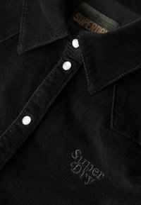Sort kordfløjlsskjorte med spidse kraver, hvide knaplukninger og et broderet "Superdry" logo på venstre foran. Tekstureret stof.