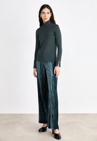 Dark green turtleneck, fitted design with long sleeves and side slits. Paired with wide-leg pleated green trousers and black shoes.
