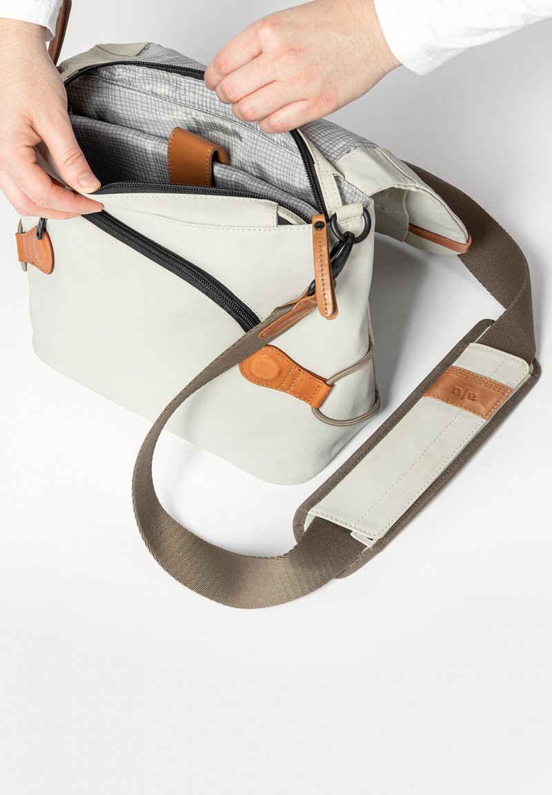 Lichtgrijze crossbodytas met een hoofdvak met ritssluiting, lederen accenten, verstelbare bruine riem en een grijs gevoerde binnenkant.