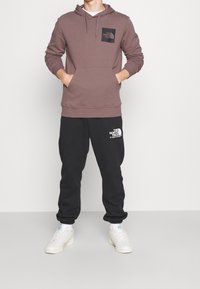 Sudadera marrón con un bolsillo frontal y un parche de logo negro, combinada con pantalones de chándal negros que presentan un estampado de logo blanco. Zapatillas blancas completan el conjunto.