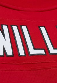 Tissu rouge avec de grandes lettres blanches en blocs, surlignées de couture noire. La texture est lisse avec des détails de couture visibles.