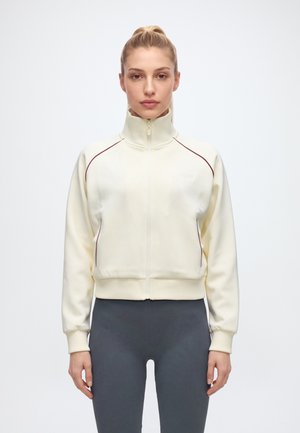 Une femme porte une veste crème zippée à col montant et passepoils bordeaux, assortie à un legging gris foncé, sur un fond uni.
