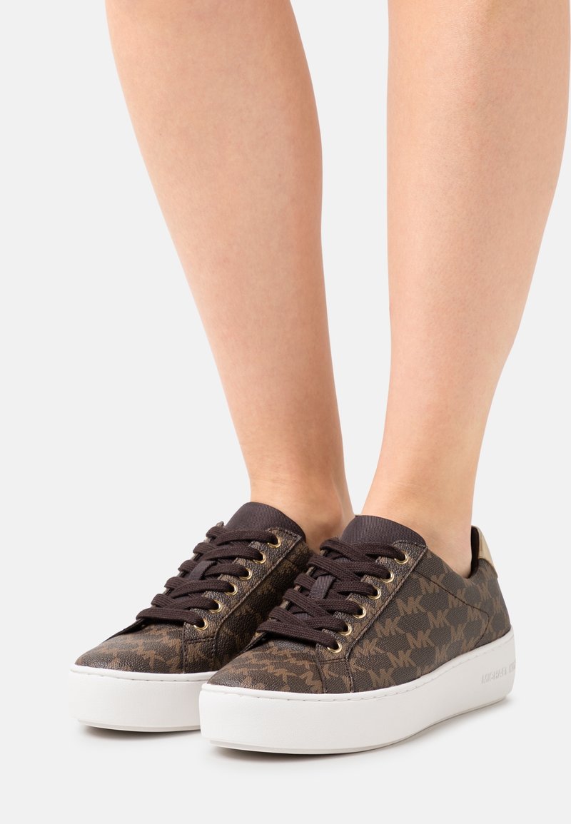 Baskets marron à motifs avec des lacets noirs et des semelles blanches épaisses portées pieds nus sur un fond clair uni.