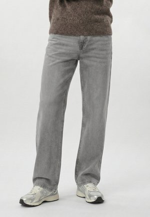 Osoba v nosených voľných sivých džínsoch a bielo-sivých teniskách stojaca na jednoduchom svetlom pozadí.