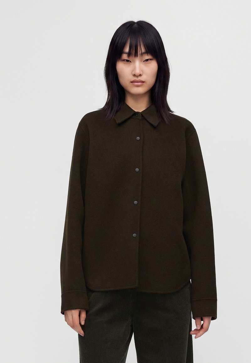 Jeune femme aux cheveux noirs et lisses porte une veste boutonnée marron foncé et un pantalon sombre sur un fond clair uni.