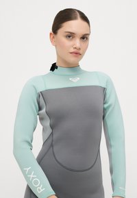 Sniegšanas kostīms ar gaiši zilu augšējo daļu un pelēku apakšējo daļu, izgatavots no gludas, elastīgas materiāla ar logotipa akcentu un augstu apkakli.