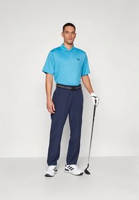Hombre de pie con un palo de golf, vistiendo una camiseta polo azul, pantalones marinos, guante de golf blanco y zapatillas deportivas blancas con rayas marinas.