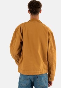 Giacca in cotone color beige con una vestibilità rilassata, dotata di colletto classico, maniche lunghe e dettagli di cucitura visibili. Indossata sopra jeans blu.