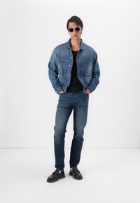 Jeansjakke i middels blå, knappet foran, med to frontlommer; kombinert med mørke jeans og svarte sko. Modell som bruker solbriller.