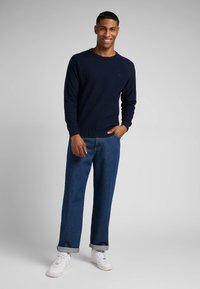Pull en maille bleu marine avec des poignets et un ourlet côtelés, associé à un jean bleu clair à jambes larges et des baskets blanches, se tenant devant un fond gris.