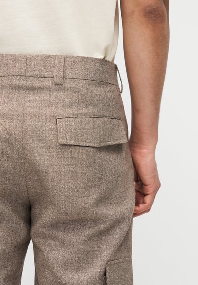 Άνδρας που φοράει μπεζ υφασμάτινο παντελόνι με τσέπη με καπάκι στο πίσω μέρος και κρεμ πουκ άμισο που έχει μπει μέσα, στέκεται μπροστά σε λευκό φόντο.