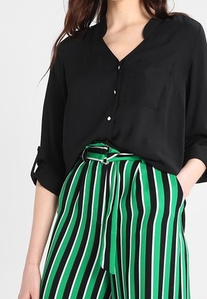 Woman wearing a black buttoned blouse with rolled sleeves tucked into high-waisted green, black, and white vertical striped pants.