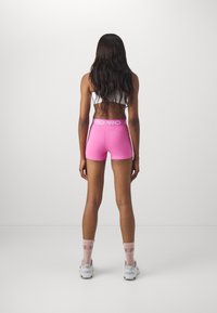 Lyserøde elastiske shorts med et hvidt logo bånd i taljen, parret med hvide sneakers og lyserøde strømper. Langt mørkt hår er synligt.