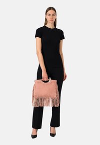 Donna in abbigliamento nero tiene in mano una borsa in suede rosa con frange e borchie metalliche, in piedi contro uno sfondo chiaro e semplice.